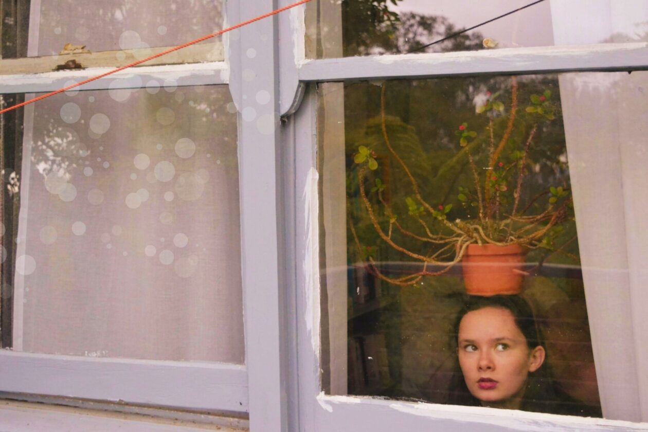 woman with a potted plant resting on her head looking out of a window with white curtains framing her