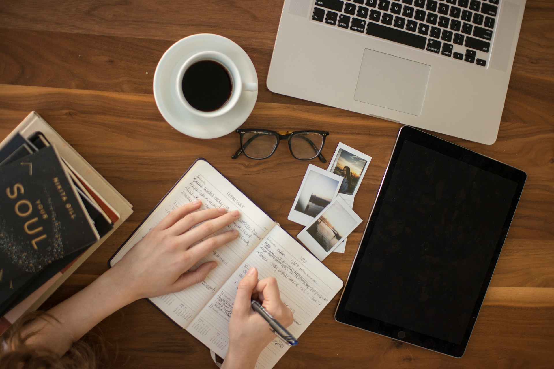 Person writing in a notebook on a wooden table. Surrounding their notebook are some books, a cup of black coffee, photographs, glasses, and a laptop.