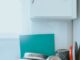clean dishes and a green cutting mat are piled onto a kitchen counter with a white tile backsplash and a white cupboard and window above them.