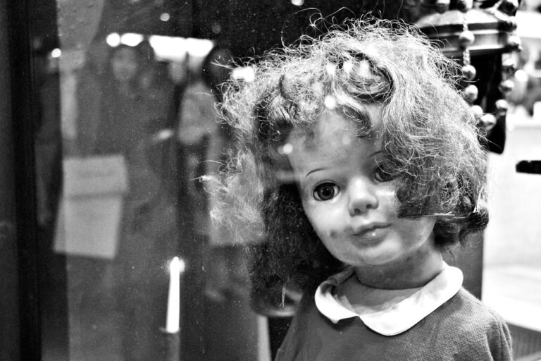 Black and white image of a vintage doll with a forlorn expression seen through glass.
