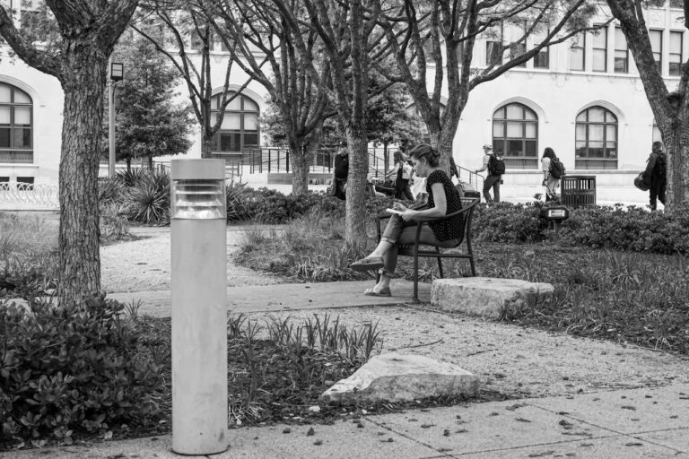 A black and white photo of an urban park with a woman reading and people walking by.