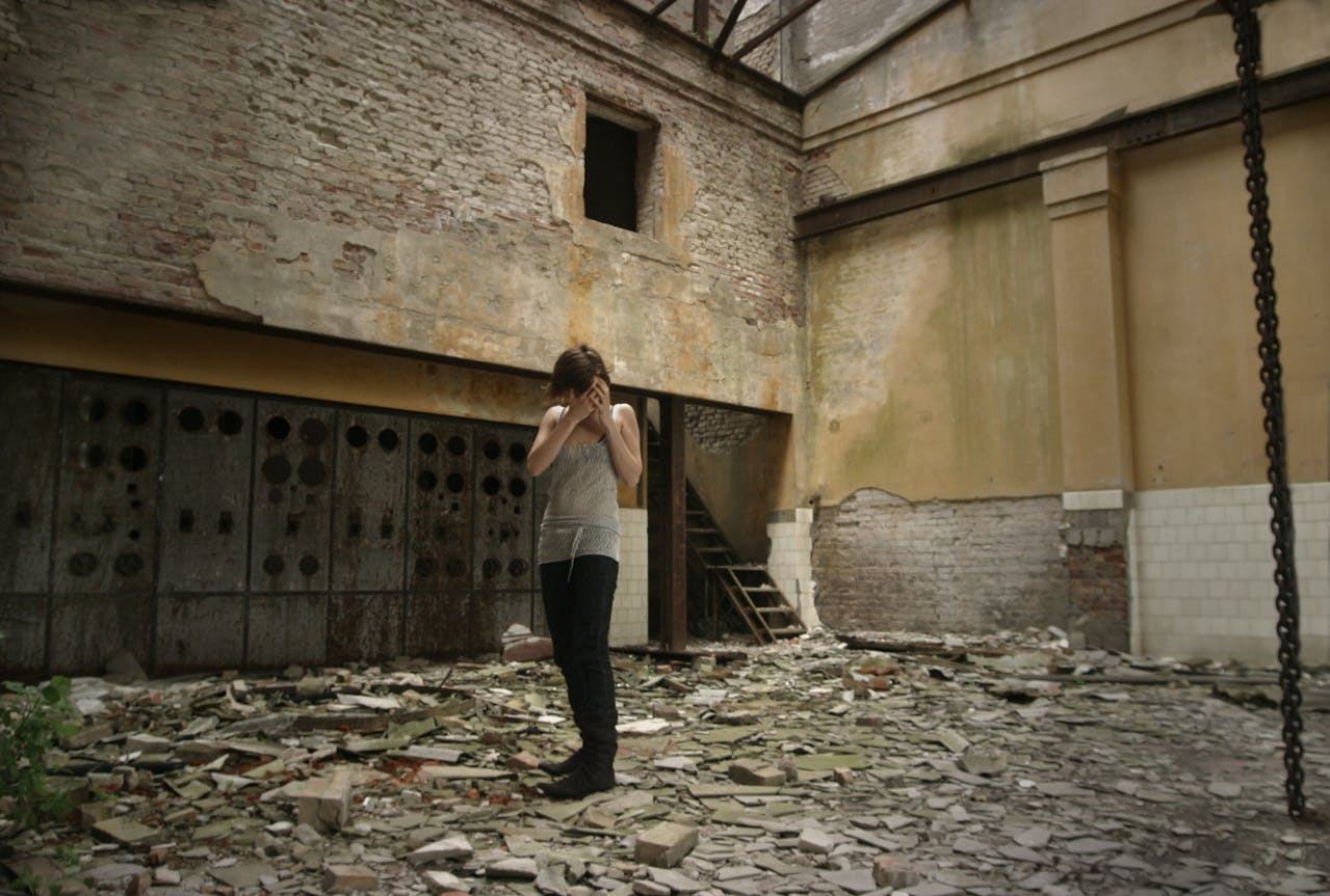 A woman stands alone in a dilapidated industrial setting, evoking solitude and decay.