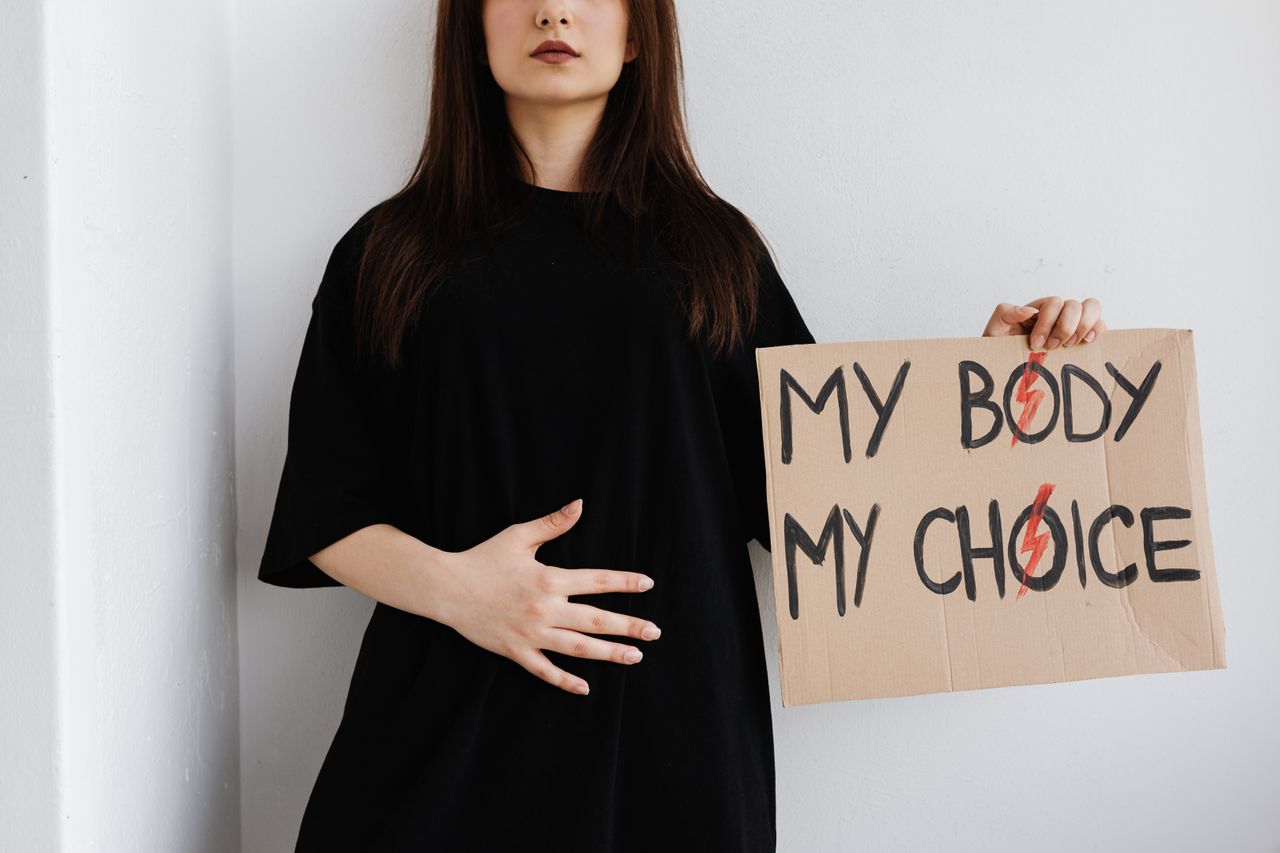 a woman wearing a black dress is standing with one hanad resting across her abdomen and the other hand holding a sign that reads 'my body my choice'